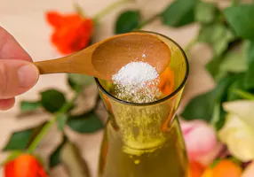 A wooden spoon with white powder is held over a glass bottle filled with liquid, surrounded by various colorful flowers in soft focus.