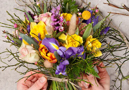 Colorful bouquet with yellow, purple, and pink flowers, held by two hands, against a light gray background, surrounded by scattered green stems and twine.