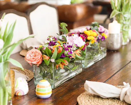 A wooden dining table is styled with colorful spring flower arrangements in rectangular glass vases, surrounded by Easter eggs, candles, and placemats