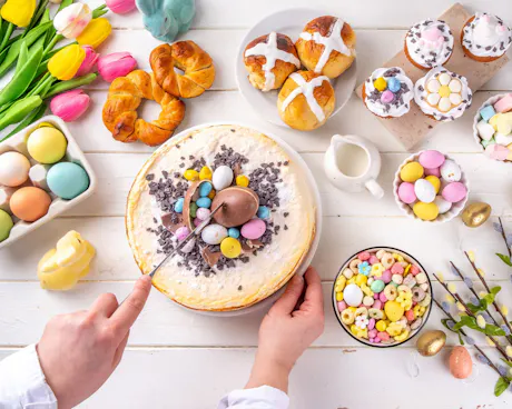 A person slices into Easter cheesecake decorated with chocolate eggs and sprinkles, surrounded by spring pastries, candies, tulips, and dyed eggs