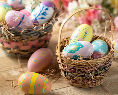 Woven Easter baskets filled with hand-painted pastel eggs rest on a rustic wood table with spring flowers and straw. Each egg is decorated with cheerful floral and spring patterns.