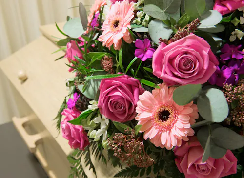 Bouquet of pink roses and daisies with green leaves rests atop a light-colored wooden casket, situated against a background of cream-colored curtains.