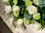 Bouquet of white roses and green carnations resting on an ornate, light-colored wooden casket. The arrangement is accented with green berries, long foliage, and eucalyptus leaves.