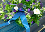 A colorful flower arrangement with white lilies, purple flowers, and a blue ribbon lies on top of a blue coffin situated in a formal setting.