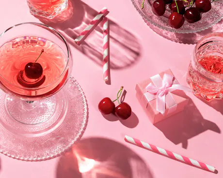 A small gift box, cherries, and charming red cocktails sit on a pink table, showing a beautiful Galentine's day setup