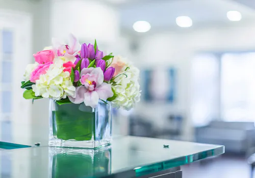 Modern floral arrangement with pink roses, purple tulips, and white hydrangea in a square glass vase, styled on a countertop.