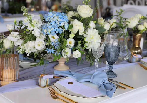 Spring table centerpiece with white and blue flowers, roses, and greenery styled for a formal celebration.