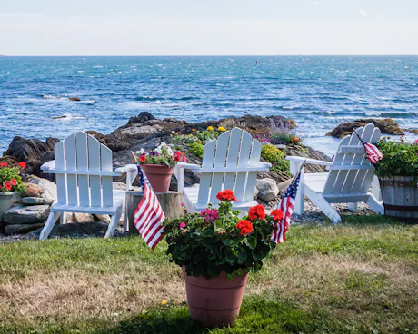 Coastal patio with American flags, red geranium plants, and white wooden chairs overlooking the ocean