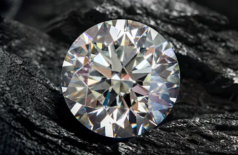 A brilliant-cut diamond resting on a rough, dark-textured surface, reflecting a spectrum of light, highlighting its facets and clarity amidst the contrast of its rugged backdrop.