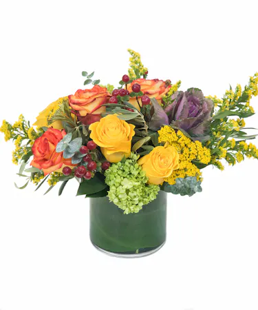 Features roses in red and orange along with red and green hypericum and green hydrangeas – skillfully designed in a cylinder vase with curly willow tips and leaves.